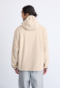 Sudadera de forro polar beige con capucha ajustable y corte relajado, que presenta una textura suave y un diseño minimalista, vista desde la parte de atrás.