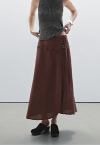 Henkilö, jolla on päällään harmaa hihaton ribbipaita, pitkä ruskea kietaisuhame, jossa on solkia, ja mustat liukuvasti puettavat puukengät, joissa on metallinappeja.