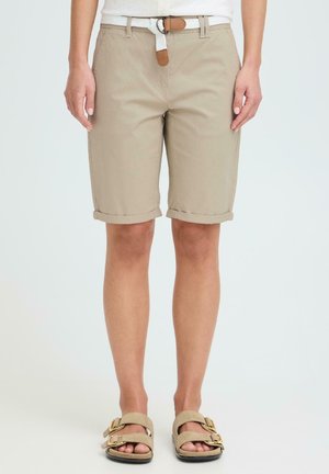 Person trägt beige knielange Shorts mit umgeschlagenen Säumen, einen weißen gewebten Gürtel und beige Sandalen mit goldenen Schnallen.