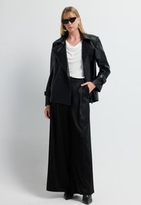 Donna che indossa una giacca di pelle nera, pantaloni larghi neri, una maglietta drappeggiata bianca, stivali neri e occhiali da sole a forma di gatto, in piedi contro uno sfondo chiaro e uniforme.