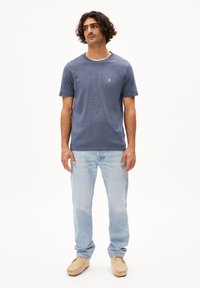 Maglietta blu navy a maniche corte con colletto rotondo, con un piccolo logo giallo. Indossata con jeans blu chiaro e scarpe tipo ciabatta beige.