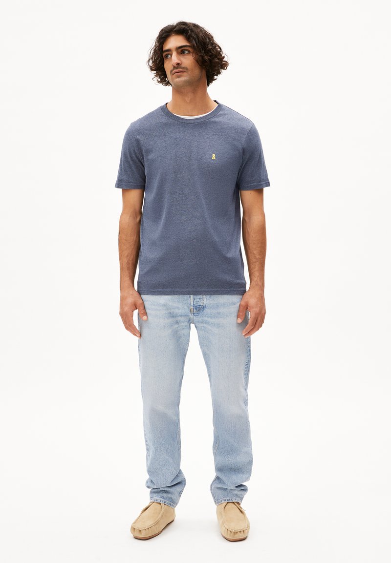 Maglietta blu navy a maniche corte con colletto rotondo, con un piccolo logo giallo. Indossata con jeans blu chiaro e scarpe tipo ciabatta beige.