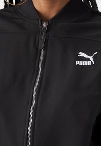 Chaqueta bomber negra con textura suave, con cremallera plateada y un logotipo blanco de Puma en el lado izquierdo. Diseño de cuello redondo.