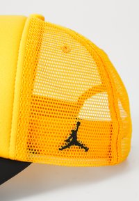 Rumena kapa z mrežastim hrbtom, s črnim logotipom Jumpman. Sprednja stran je enobarvno rumena, medtem ko imata stranski del teksturiran mrežast dizajn.