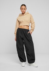 Felpa beige tagliata con cappuccio regolabile, abbinata a pantaloni neri larghi. Modello che indossa sneaker bianche, design minimalista.