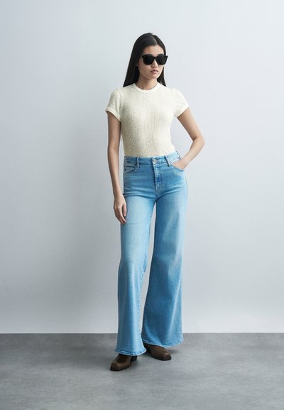 Γυναίκα με μαύρα γυαλιά ηλίου, κρεμ πλεκτή μπλούζα με κοντά μανίκια, ανοιχτό μπλε ψηλόμεσα καμπανες τζιν και καφέ μπότες, στέκεται με το ένα χέρι στην τσέπη.