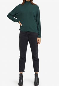 Maglia a maniche lunghe verde scuro con collo a camicia, caratterizzata da una vestibilità rilassata e spacco laterale, abbinata a jeans neri con risvolto e stivali neri alla caviglia.