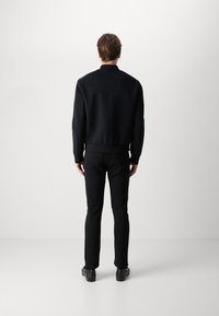 Veste bomber noire avec poignets et ourlet côtelés, associée à un jean noir ajusté et des chaussures noires, vue de dos. Texture de tissu lisse.