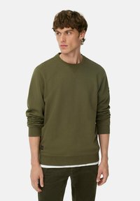 Olijfgroene crewneck sweatshirt van zachte stof met ribbels aan de mouwen, voorzien van een subtiele logopatch aan de onderrand. Gestyled met rechte broek.