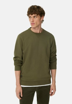 Olivgrüner Rundhalsausschnitt-Pullover aus weicher Stoffqualität mit gerippten Bündchen, versehen mit einem dezenten Logo-Patch am unteren Saum. Kombiniert mit gerade geschnittenen Hosen.
