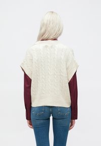 Pleteni pulover brez rokavov v kremni barvi z kratkimi rokavi, v kombinaciji z maroon majico z dolgimi rokavi in modrimi kavbojkami, prikazan s hrbtne strani.