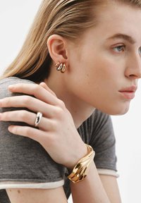 Pendientes de metal dorado con un diseño curvado, una pulsera de brazalete dorada a juego y un anillo plateado elegante contra un fondo de camisa gris.