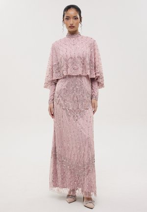 Donna che indossa un lungo abito rosa con intricati ricami di perline e un mantello abbinato, in piedi davanti a uno sfondo semplice e chiaro.