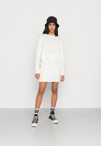 Weißer gestrickter, kurzer Pullover, kombiniert mit einem passendem, gestrickten Rock. Ein schwarzer Bucket Hat, schwarze High-Top Sneakers und gestreifte Socken runden das Outfit ab.