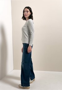 Jasnoszary prążkowany top z długim rękawem w połączeniu z ciemnoniebieskimi jeansami o szerokich nogawkach i beżowymi trampkami; modelka stoi na tle jednolitego tła.
