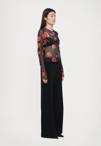 Black sheer long-sleeve top with red floral print and a scalloped hem, paired with high-waisted black wide-leg trousers.