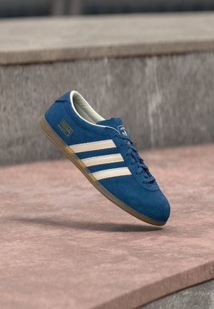 Kék suede sportcipő krémszínű csíkokkal és fűzőkkel, gumi talppal, texturált felülettel, lekerekített orral és oldalán márkajelzéssel.