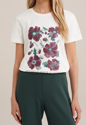 White short-sleeve t-shirt with large purple floral print and French text, worn with high-waisted dark green pants.