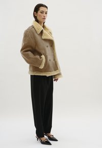 Donna in una giacca di shearling beige sopra pantaloni neri a gamba larga e scarpe col tacco a punta nere, in un contesto con sfondo chiaro.