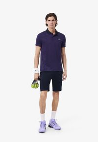 Lila texturerad polo skjorta med svart krage, i kombination med marinblå shorts. Modellen håller en meshväska med gröna tennisbollar och bär lavendelfärgade skor.