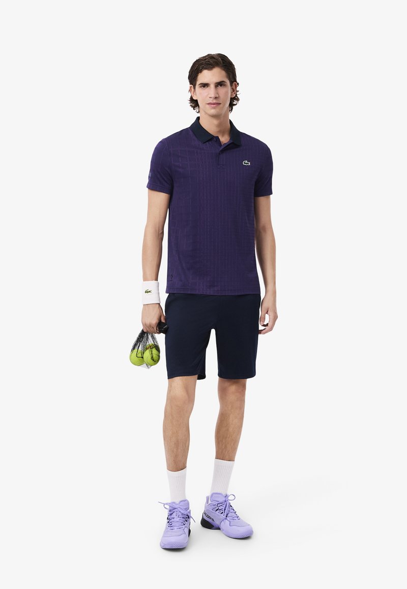 Polo shirt viola con texture e colletto nero, abbinato a pantaloni corti blu scuro. Il modello tiene in mano una borsa a rete con palline da tennis verdi e indossa scarpe color lavanda.