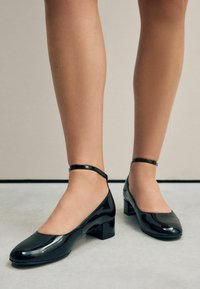 Schwarze Lacklederschuhe mit quadratischer Zehenpartie und kleinem Blockabsatz. Verfügt über einen Knöchelriemen für einen sicheren Sitz. Glatte, glänzende Oberfläche.