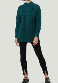 Tealowy sweter dziany z wzorami w warkocz, wysoki kołnierz i ściągacz u dołu, w połączeniu z czarnymi legginsami z bocznymi rozcięciami i błyszczącymi czarnymi butami.