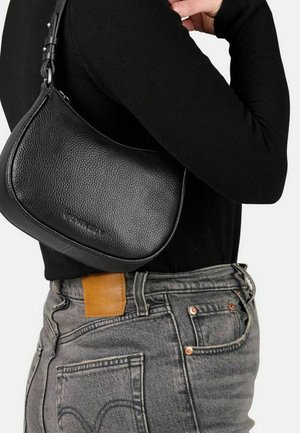 Persona che indossa un top nero a maniche lunghe e jeans grigi, con una piccola borsa a tracolla in pelle texturizzata nera al suo fianco.