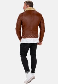 Giubbotto di pelle marrone con colletto in shearling, schiena inferiore imbottita, maniche aderenti e finitura liscia. Indossato sopra jeans neri e scarpe bianche.