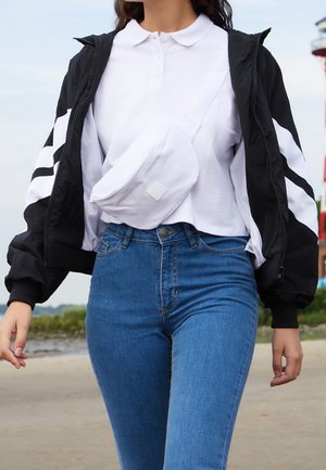 Femme marchant en plein air, portant un jean bleu, un polo blanc, une veste noire et blanche, et un sac bandoulière blanc, sur un fond naturel flou.