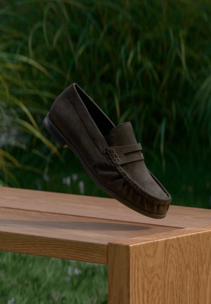 Brauner Wildleder-Loafer, der über einer Holzbank schwebt, mit grünem Gras und Pflanzen im Hintergrund unscharf.