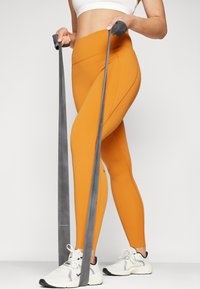 Oransje tights laget av elastisk stoff, med høy midje og sidesømmer, kombinert med grå motstandsbånd og hvite treningssko.