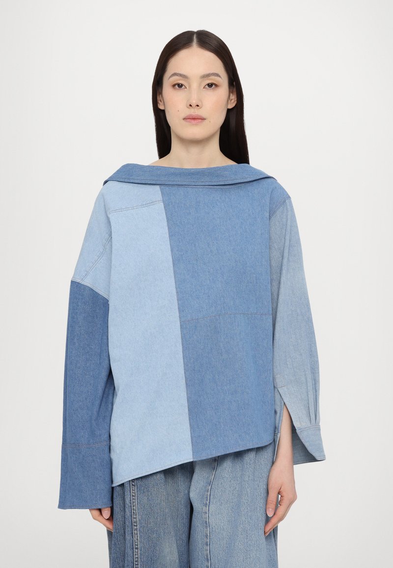 Denim sweater in varying shades of blue, featuring a wide collar, oversized sleeves, and a boxy cut with visible seam details.