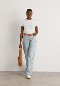 Hvit t-skjorte med blå tekst, lyseblå høytlivte jeans, brun belte, og en brun skulderveske. Hvite sneakers fullfører antrekket.
