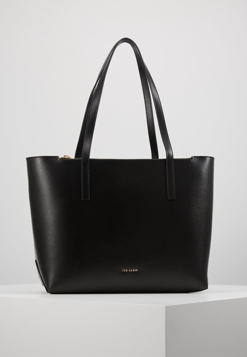 Sac fourre-tout en cuir noir avec doubles bretelles, forme rectangulaire et petit logo doré « Ted Baker » à l'avant, sur un fond uni.