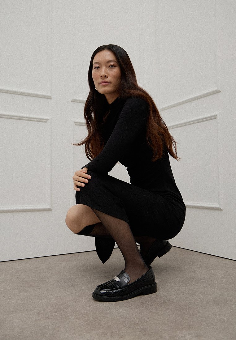 Schwarzes knielanges Kleid mit langen Ärmeln, schwarze blickdichte Strumpfhosen und glänzende schwarze Slipper mit gestepptem Muster und silbernen Hardware-Details.