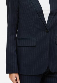 Blazer marine à fines rayures avec fermeture à un bouton et poches à rabat porté sur un haut blanc, assorti à un pantalon à fines rayures.