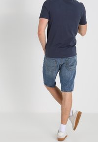 La camiseta de manga corta azul marino combinada con pantalones cortos de mezclilla azul deslavado, llevada con calcetas blancas y zapatos casuales color beige, vista desde atrás.