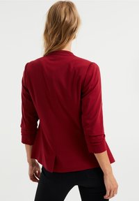 Blazer rojo ajustado con mangas de tres cuartos, textura suave, espalda entallada y un sutil efecto peplum en el dobladillo.