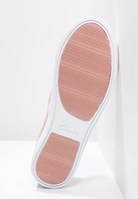 Podešev růžové a bílé tenisky s texturovaným gripem a logem "Clarks", nakloněná proti bílé rohové zdi a podlaze.