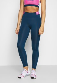 Vysoké legíny v tmavě modré barvě, vyrobené z hladkého materiálu a s těsným střihem. V kombinaci s bílými a růžovými sportovními botami se vzorem.