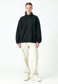 Veste en polaire noire avec fermeture éclair à l'avant, col montant et poches latérales ; portée avec un pantalon cargo crème et des baskets blanches. Coupe texturée et oversize.