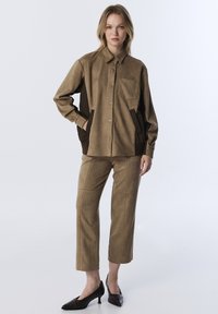 Vrouw die staat in een tan suède shirtjack met donkere zijkanten, bijpassende cropped pantalons en zwarte lage hak schoenen op een witte achtergrond.