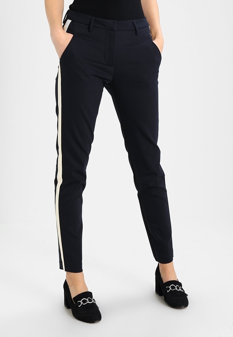 Femme portant un pantalon noir slim avec des bandes blanches sur les côtés, mains dans les poches, associé à des mocassins noirs à talons blocs avec détail chaîne argentée.