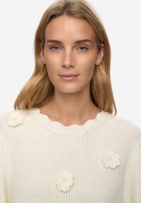 Krémový pletený svetr s vroubkovaným výstřihem a třemi texturovanými květinovými aplikacemi. Má volný střih a měkký, prodyšný materiál.