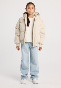 Ragazzina in piedi che indossa una giacca a vento beige, una maglietta bianca, jeans larghi azzurri chiari e sneakers bianche su uno sfondo neutro.