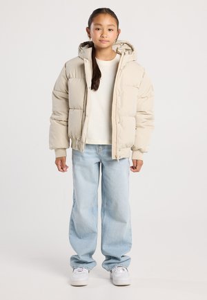 Une jeune fille se tient debout, portant une doudoune beige, une chemise blanche, un jean large bleu clair et des baskets blanches sur un fond uni.
