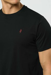 Vyras dėvi paprastą juodą marškinėlį su apvalia iškirpte ir mažą raudoną siuvinėto logotipo ant kairiojo krūtinės.