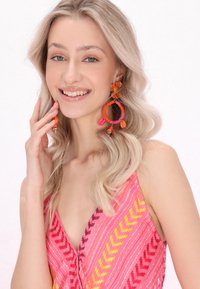 Barevné korálkové náušnice mají kulatý design s akcenty v růžové, oranžové a fialové barvě. Modelka má na sobě živou růžovou halenku se vzorem chevron.