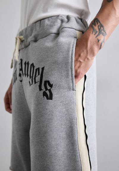 Close-up of a person wearing grey sweatpants with "Palm Angels" text, a side cream stripe, and a visible tattoo on the wrist.